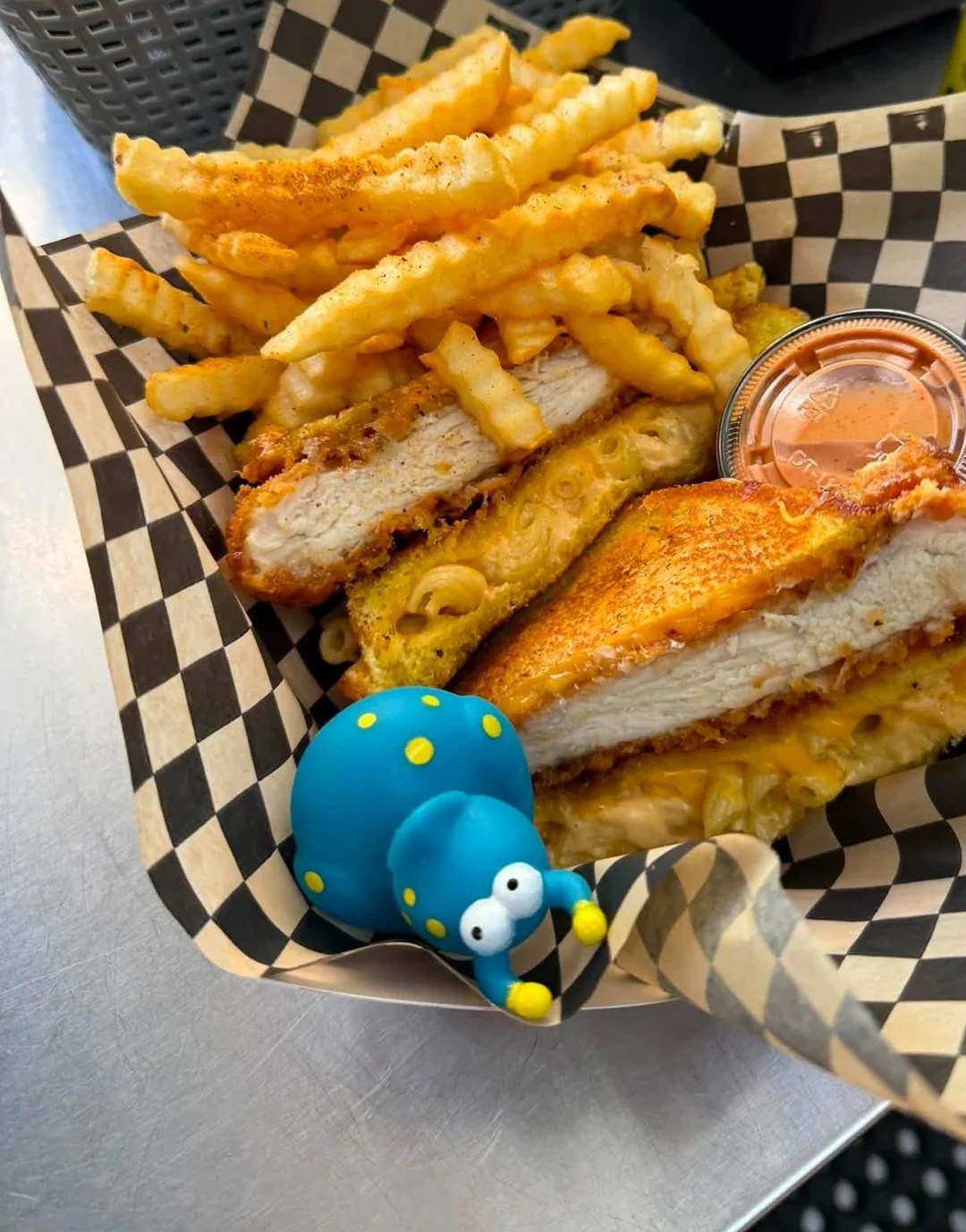 Crispy chicken sandwich with macaroni and cheese, seasoned crinkle fries, dipping sauce, and a small blue toy on a checkered food liner.