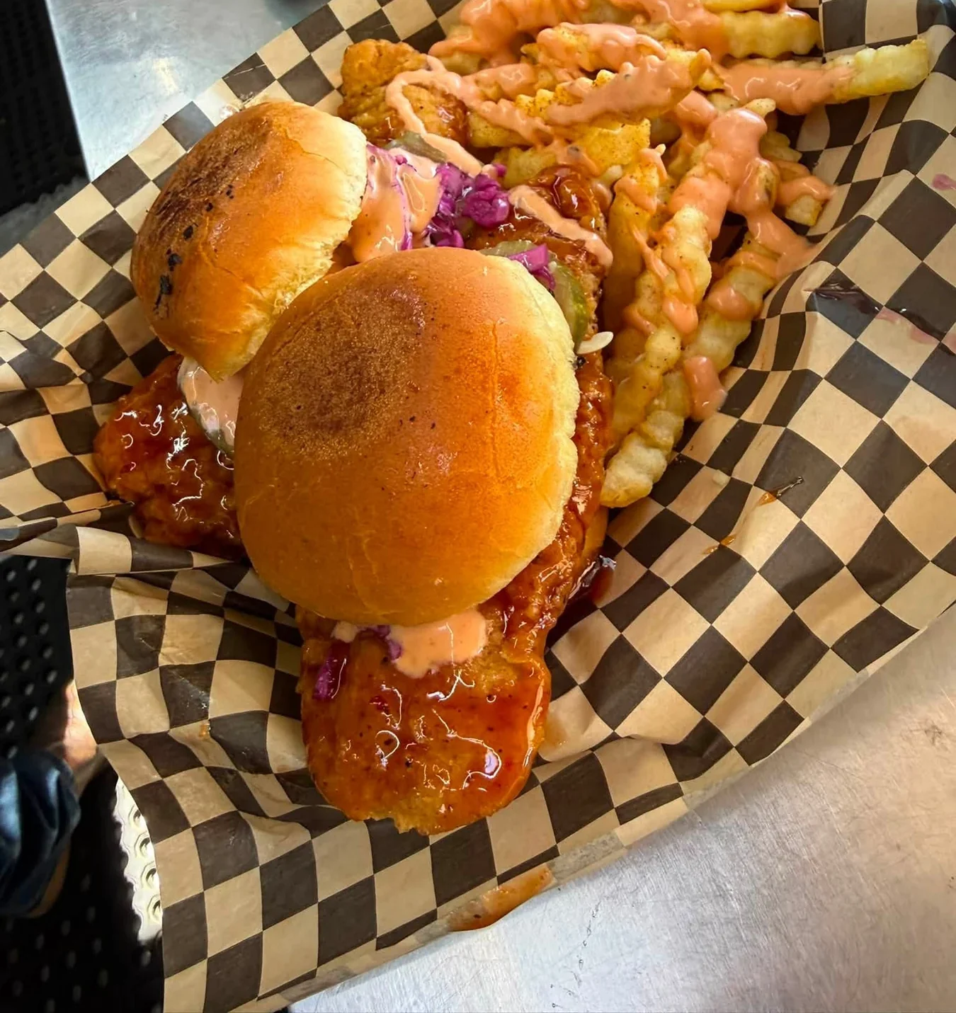 Two chicken sandwiches with slaw and sauce beside crinkle fries drizzled with sauce on a checkered food liner.