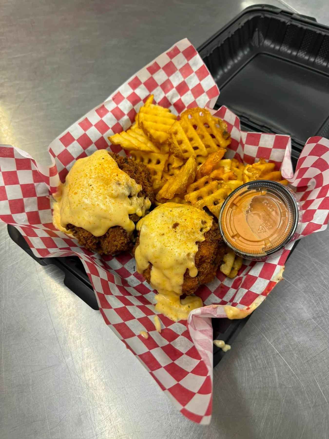 Two pieces of fried chicken topped with cheese sauce beside seasoned waffle fries and a dipping sauce.