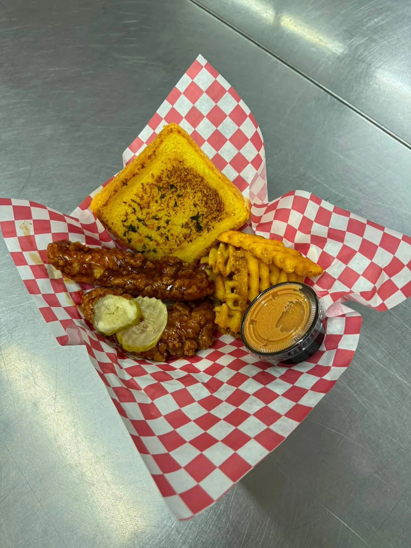 Sauced chicken tenders with pickles, waffle fries, toasted bread, and dipping sauce on a red checkered liner.