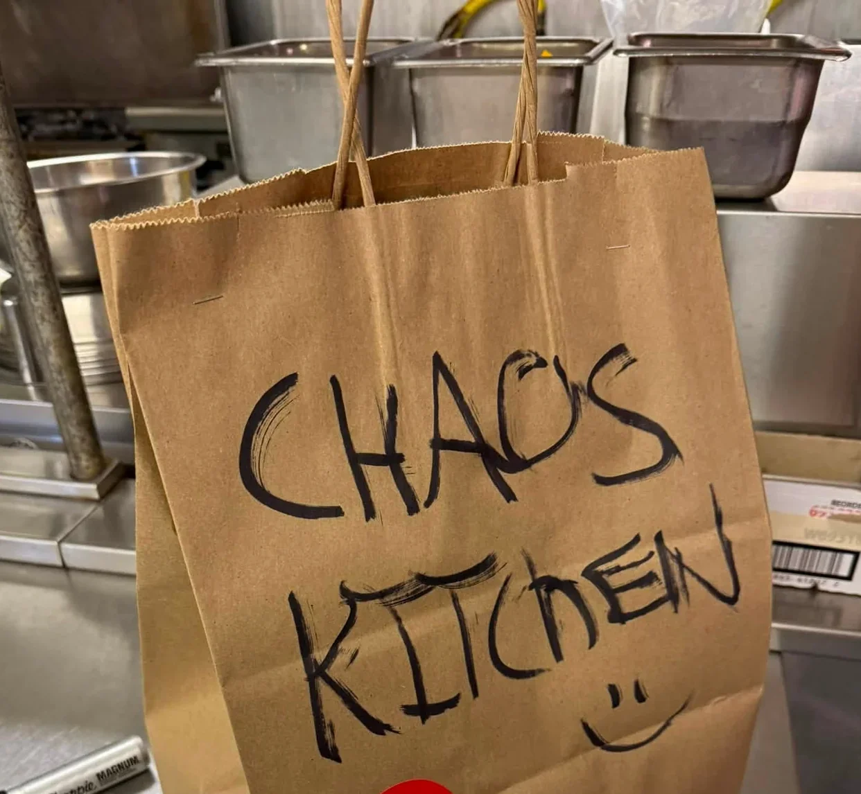 Brown takeout bag hand-lettered โCHAOS KITCHENโ with a smiley face, photographed in a commercial kitchen.